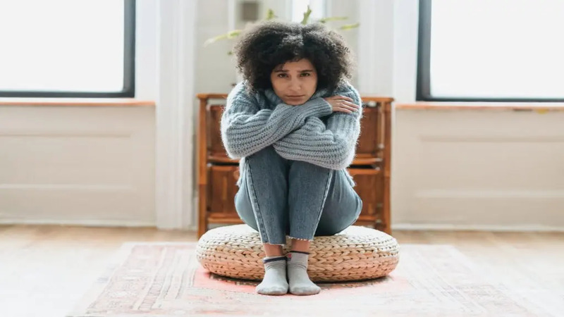 A person sitting cross-legged on a round cushion in a room with two windows. They are wearing a gray sweater, blue jeans, and gray socks.